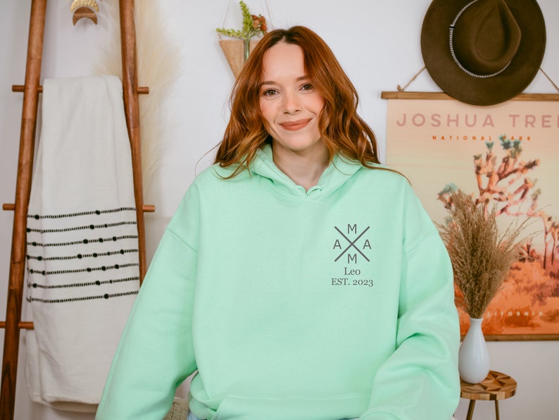 Peut inclure: Un sweat &agrave; capuche vert clair avec un logo noir et blanc qui dit "MAMA" avec une croix au milieu et "Leo EST. 2023" en dessous.