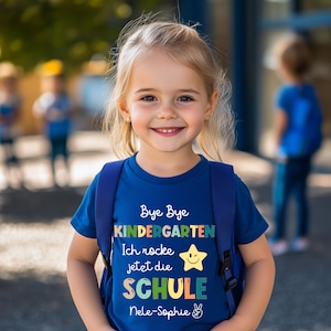 Puede incluir: Una ni&ntilde;a peque&ntilde;a lleva una camiseta azul con una mochila. La camiseta tiene el texto "Bye Bye Kindergarten" en letras coloridas, junto con las palabras "Ich rocke jetzt die SCHULE" y un gr&aacute;fico de estrella sonriente. La ni&ntilde;a sonr&iacute;e a la c&aacute;mara.