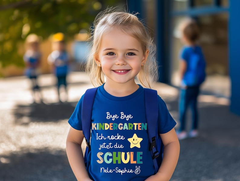 Puede incluir: Una ni&ntilde;a peque&ntilde;a lleva una camiseta azul con una mochila. La camiseta tiene el texto "Bye Bye Kindergarten" en letras coloridas, junto con las palabras "Ich rocke jetzt die SCHULE" y un gr&aacute;fico de estrella sonriente. La ni&ntilde;a sonr&iacute;e a la c&aacute;mara.