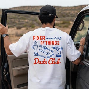 Könnte beinhalten: Weißes T-Shirt mit einem rot-blauen Grafikdesign. Das Design enthält die Worte "FIXER knower of stuff OF THINGS" und "Dads Club" sowie Illustrationen von Werkzeugen und einer Kaffeetasse. Die Person steht neben einem Fahrzeug.