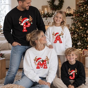 Op de afbeelding: Een collectie zwarte en witte sweaters met ronde hals, voorzien van een grote rode letter met witte sneeuwvlokpatronen, bekroond met een kerstmuts en rendiergeweien. De letters zijn gepersonaliseerd met de namen Papa, Mama, Anna en Jonas.