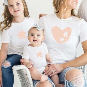 May include: A family of three wearing white t-shirts with peach-colored puzzle piece designs. The adult shirt features a heart with two puzzle pieces inside. The child's shirt has a single puzzle piece. The family is sitting together, with the baby sitting on the adult's lap.
