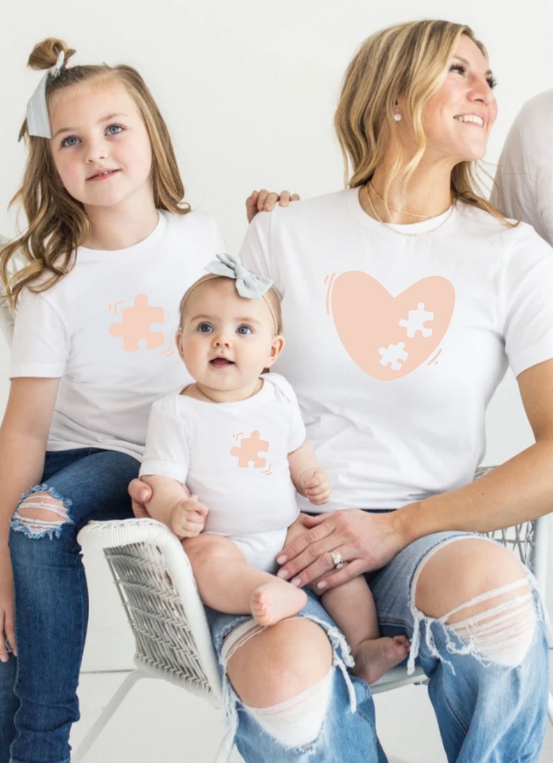 May include: A family of three wearing white t-shirts with peach-colored puzzle piece designs. The adult shirt features a heart with two puzzle pieces inside. The child's shirt has a single puzzle piece. The family is sitting together, with the baby sitting on the adult's lap.