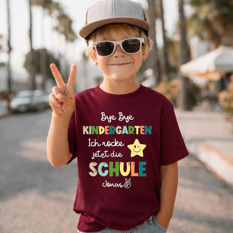 Puede incluir: Una camiseta burdeos con el texto "Bye Bye Kindergarten Ich rocke jetzt die SCHULE Jonas" en letras coloridas. La camiseta presenta un gr&aacute;fico de estrella sonriente. El ni&ntilde;o lleva una gorra de camionero gris y gafas de sol.