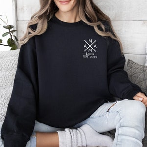 Peut inclure: Sweat-shirt noir avec un motif graphique blanc repr&eacute;sentant un monogramme avec les lettres "MAMA" et le texte "Louis EST. 2023".