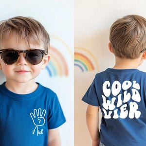 Pode incluir: Uma criança usando uma camiseta azul marinho e óculos de sol. A frente da camisa apresenta um desenho de mão e o nome "Nick". A parte de trás da camisa exibe as palavras "Cool Wild Vier" em letras brancas.