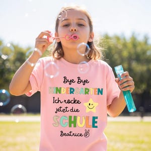 Puede incluir: Una persona joven lleva una camiseta rosa claro con las palabras "Bye Bye Kindergarten" e "Ich rocke jetzt die SCHULE" impresas. La camiseta tambi&eacute;n presenta una estrella gui&ntilde;ando un ojo y el nombre "Beatrice". La persona est&aacute; soplando burbujas.