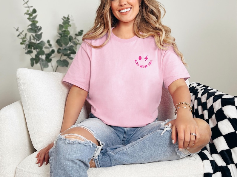 Peut inclure: Une femme portant un t-shirt rose avec le texte "Girl Mom Era" et un smiley rose avec un &eacute;clair. Elle est assise sur un canap&eacute; blanc portant un jean bleu.