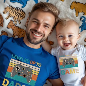 以下が含まれることがあります： 青いTシャツと、お揃いのゲームコントローラーのグラフィックが入った白いボディスーツ。青いシャツには「LEVELED UP TO DAD」と書かれており、ボディスーツには「PLAYER 2 HAS ENTERED GAME」と書かれています。どちらもレトロな配色です。