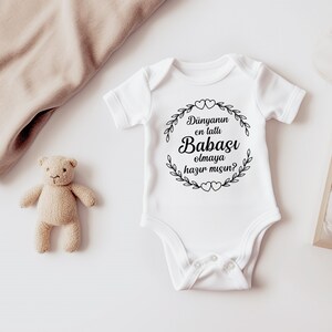 May include: White baby onesie with black text that reads "Dunyannun en tatlu Babasi olmaya hazir misin?"  The text is inside a wreath with hearts.