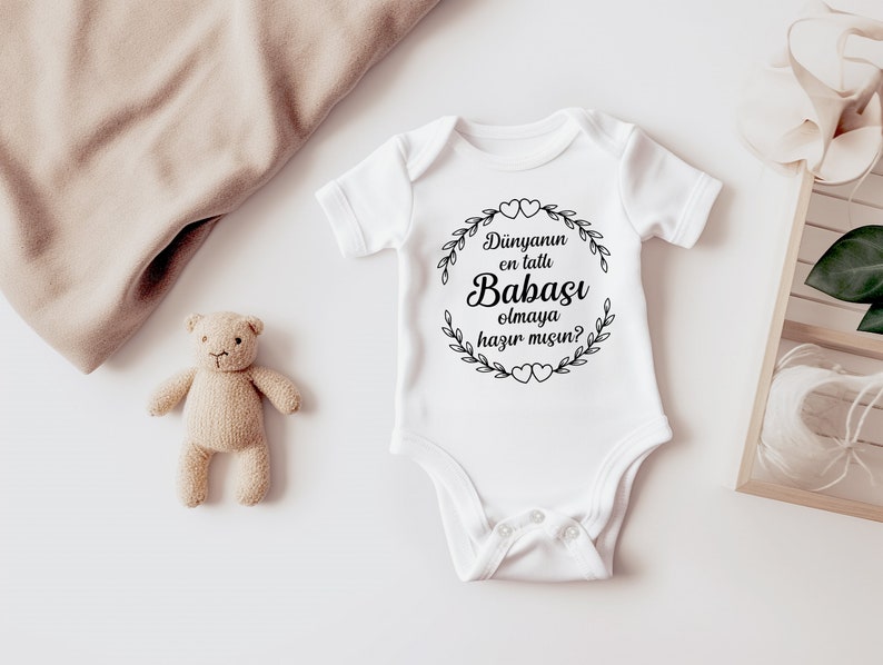 May include: White baby onesie with black text that reads "Dunyannun en tatlu Babasi olmaya hazir misin?"  The text is inside a wreath with hearts.