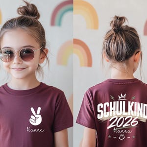 May include: A maroon short-sleeved t-shirt featuring a white peace sign and the name Hanna on the front. The back displays the text SCHULKIND 2026, a crown icon, the name Hanna, and a smiley face graphic with stars in white and beige tones.
