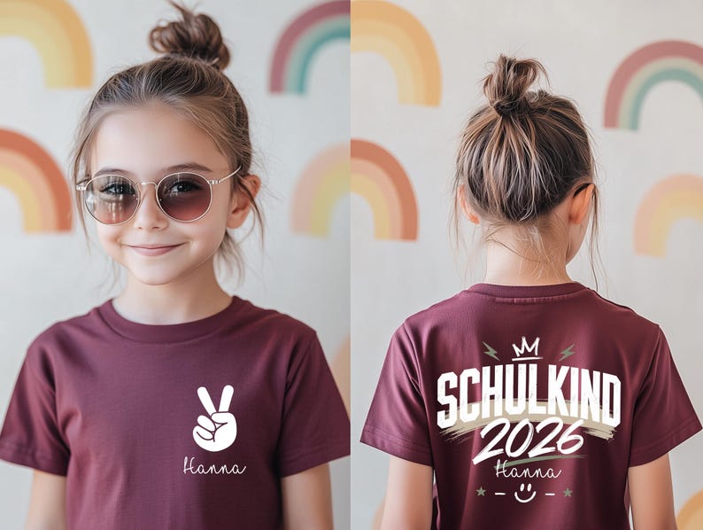May include: A maroon short-sleeved t-shirt featuring a white peace sign and the name Hanna on the front. The back displays the text SCHULKIND 2026, a crown icon, the name Hanna, and a smiley face graphic with stars in white and beige tones.