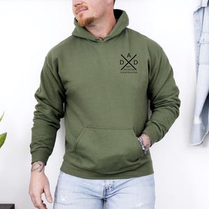 May include: Olive green hoodie with a front pocket and a hood. The hoodie features a black graphic on the chest that says "DAD" with crossed arrows and the text "EST. 2021/24 Love & Kisses & Y'all".