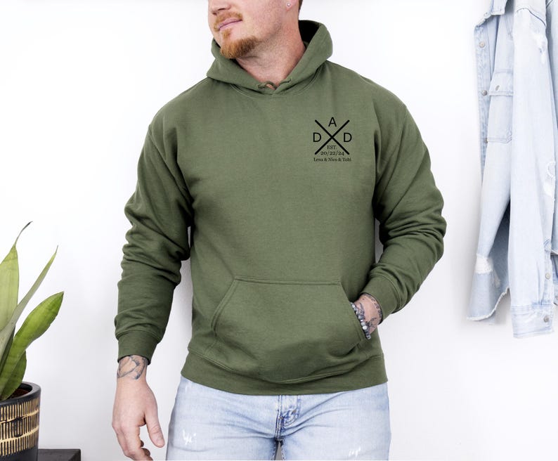 May include: Olive green hoodie with a front pocket and a hood. The hoodie features a black graphic on the chest that says "DAD" with crossed arrows and the text "EST. 2021/24 Love & Kisses & Y'all".