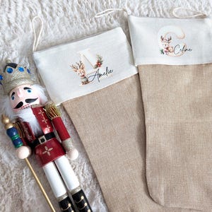 Op de afbeelding: Twee beige kerstsokken met gepersonaliseerde namen en rendierillustraties. Eén sok heeft de naam "Amelie" en de andere "Celine". Een decoratieve notenkraker staat in de buurt en voegt een feestelijk tintje toe aan de scène.