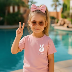 May include: A light pink short-sleeved t-shirt featuring a white graphic of a hand making a peace sign and the name Hanna printed in white script below it. The shirt has a crew neckline and is made of a solid, plain cotton-like fabric.