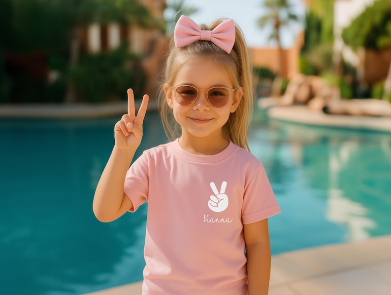 May include: A light pink short-sleeved t-shirt featuring a white graphic of a hand making a peace sign and the name Hanna printed in white script below it. The shirt has a crew neckline and is made of a solid, plain cotton-like fabric.
