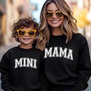 May include: A black sweatshirt with the word "MAMA" printed in white on the front. A child is wearing a black sweatshirt with the word "MINI" printed in white on the front. Both are wearing yellow sunglasses.