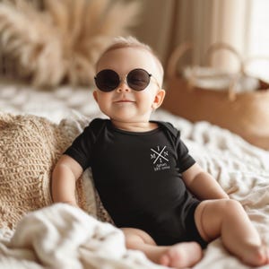 Pu&ograve; includere: Un bambino che indossa un body nero con una grafica bianca che dice "MINI" con una croce e il testo "Anton EST. 2024". Il bambino indossa occhiali da sole neri e siede su un letto.