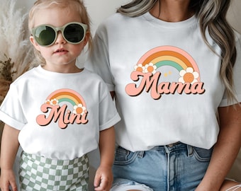 May include: White t-shirts with rainbow and floral designs. One shirt says "Mama" in a retro font, and the other says "Mini". The shirts are worn by a mother and child. The child is wearing green sunglasses and checkered shorts.