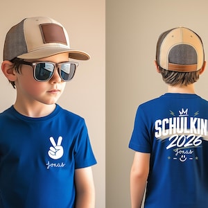 May include: A blue t-shirt featuring a white peace sign graphic and the name Jonas on the front, with the text SCHULKIND 2026, a crown, and a smiley face on the back. The outfit includes a beige and brown mesh trucker hat and dark sunglasses.