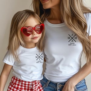 Pu&ograve; includere: Due magliette bianche con un design grafico nero. La maglietta pi&ugrave; grande ha il testo "A M A M Lucie EST. 2021". La maglietta pi&ugrave; piccola ha il testo "M I N I Lucie EST. 2021".