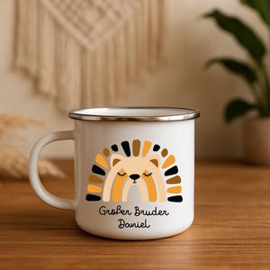 May include: A white enamel mug with a silver rim featuring a graphic of a stylized lion with a rainbow-shaped mane in shades of yellow, brown, and black. Below the lion, the text reads Gro&szlig;er Bruder Daniel in a black, rounded script font.