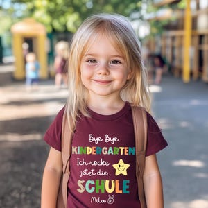 Puede incluir: Una ni&ntilde;a rubia sonr&iacute;e, vistiendo una camiseta granate con el texto "Bye Bye Kindergarten, Ich rocke jetzt die SCHULE" y una estrella. Lleva una mochila marr&oacute;n y est&aacute; de pie frente a una escuela.