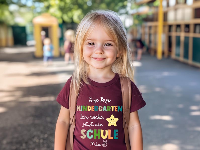Puede incluir: Una ni&ntilde;a rubia sonr&iacute;e, vistiendo una camiseta granate con el texto "Bye Bye Kindergarten, Ich rocke jetzt die SCHULE" y una estrella. Lleva una mochila marr&oacute;n y est&aacute; de pie frente a una escuela.