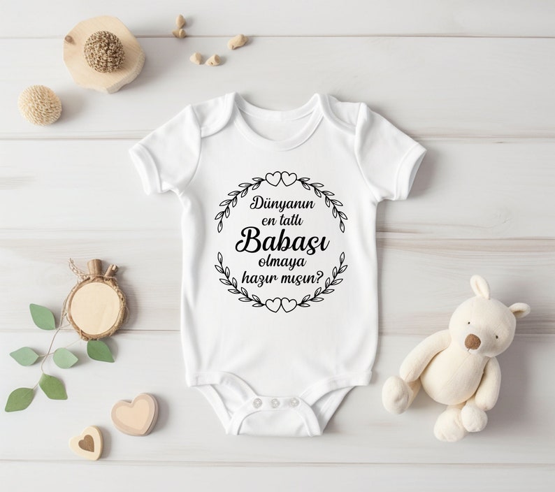 May include: White baby onesie with black text that reads "D&uuml;nyanın en tatlı Babası olmaya hazır mısın?"  The text is surrounded by a wreath of leaves and hearts.