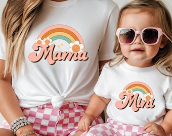 May include: A white t-shirt with a rainbow and flowers graphic and the text "Mama" in a retro font. A second white t-shirt with the same rainbow and flower graphic and the text "Mini" in a retro font.