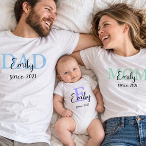 Könnte beinhalten: Zwei weiße T-Shirts und ein weißer Baby-Body. Ein T-Shirt zeigt den hellblauen Text DAD mit Emily since 2021. Das andere T-Shirt zeigt den hellgrünen Text MOM mit Emily since 2021. Der Body zeigt den violetten Buchstaben E mit Emily since 2021.