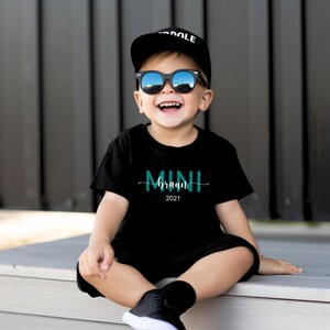 May include: A young child wearing a black t-shirt with the text "MINI braun 2021" in teal blue lettering. The child is wearing a black baseball cap with the word "DOLE" in white lettering and black sunglasses.