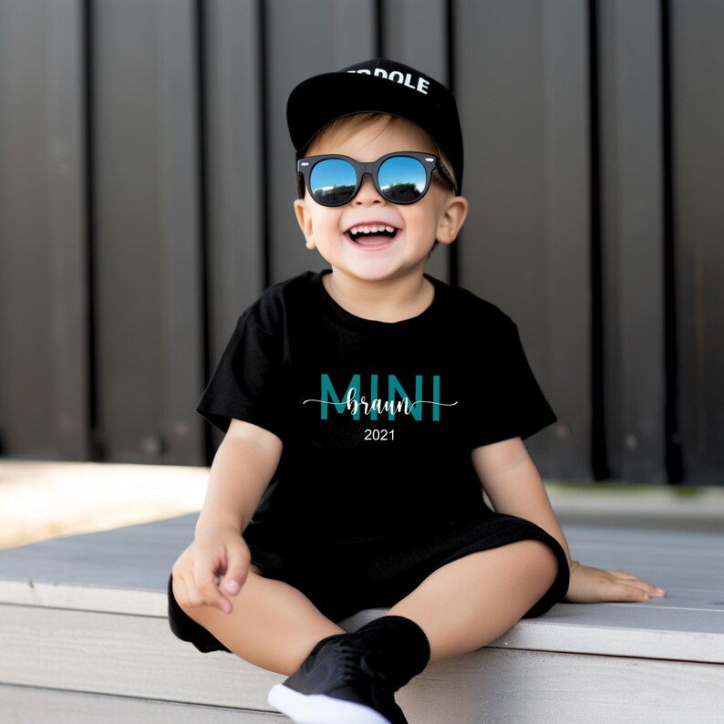 May include: A young child wearing a black t-shirt with the text "MINI braun 2021" in teal blue lettering. The child is wearing a black baseball cap with the word "DOLE" in white lettering and black sunglasses.