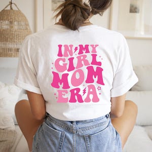 Peut inclure: T-shirt blanc avec un motif rose et blanc qui dit "In My Girl Mom Era" avec un &eacute;clair et des &eacute;toiles.