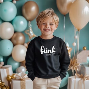 May include: A young boy wearing a black sweatshirt with the text "fünf LEOPOLD" in white. He is standing in front of a blue background with balloons and wrapped gifts.