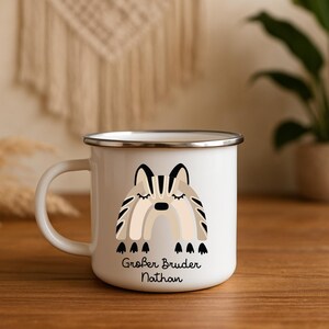 May include: A white enamel mug with a silver rim and a handle on the left side. The front features a graphic of a stylized rainbow in shades of beige, brown, and black, with the German text "Gro&szlig;er Bruder Nathan" printed in black below it.