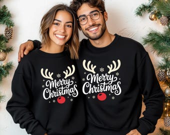 May include: Two people wearing matching black sweatshirts with a festive Christmas design. The design includes reindeer antlers, snowflakes, the words "Merry Christmas," and a red nose. The sweatshirts are a cozy and stylish choice for the holiday season.