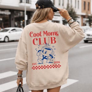 以下が含まれることがあります： 赤色の「Cool Moms CLUB」の文字と青い漫画のコーヒーカップのグラフィックが特徴のベージュのスウェットシャツ。デザインには「EST. 2026」の文字と、下部に赤と白のチェック柄が含まれています。