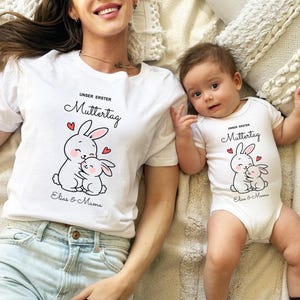 Puede incluir: Conjunto de camiseta blanca y body para beb&eacute;, con un dise&ntilde;o de conejito de dibujos animados y el texto "UNSER ERSTER Muttertag", as&iacute; como los nombres "Elias & Mama". El dise&ntilde;o incluye corazones rojos. El adulto lleva una camiseta, el beb&eacute; un body.