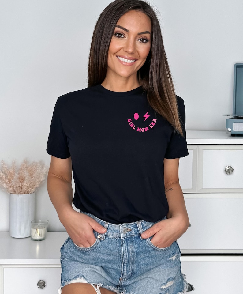 Peut inclure: Un t-shirt noir avec un graphique rose d'un visage souriant avec un &eacute;clair et le texte "Girl Mom Era".