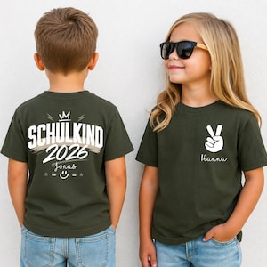 May include: Two olive green t-shirts. The back of one shirt features the text SCHULKIND 2026, a crown, a smiley face, and the name Jonas. The front of the other shirt features a white peace sign graphic and the name Hanna in white script.