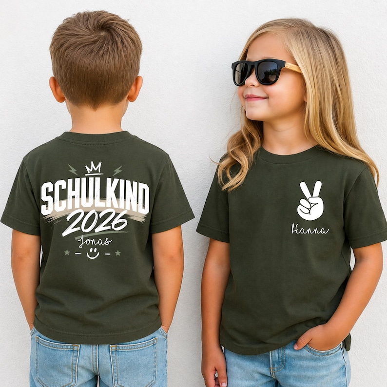 May include: Two olive green t-shirts. The back of one shirt features the text SCHULKIND 2026, a crown, a smiley face, and the name Jonas. The front of the other shirt features a white peace sign graphic and the name Hanna in white script.