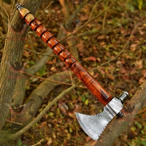 May include: A hand-forged tomahawk with a Damascus steel head and a wooden handle. The handle is made of dark wood with a reddish hue and has a decorative pattern. The head of the tomahawk has a unique pattern and is polished to a high shine.