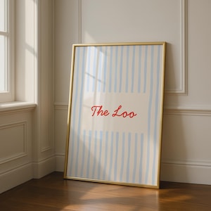 Può includere: Una stampa artistica rettangolare in una sottile cornice dorata con sfondo color crema e strisce verticali azzurre. Al centro della stampa è presente la scritta The Loo in un carattere rosso, corsivo e in stile calligrafico.