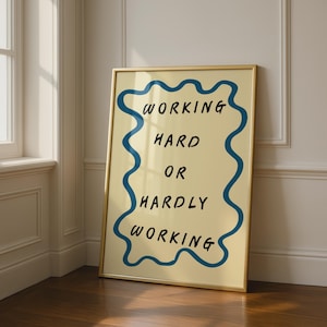 May include: A rectangular wall art print in a thin gold frame featuring a cream-coloured background with a wavy blue border. The black text in the centre reads: WORKING HARD OR HARDLY WORKING. The print is displayed in a vertical orientation.