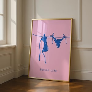 May include: A rectangular wall art print in a gold frame featuring a pink background with a dark blue line drawing of a bikini hanging on a clothesline. The text Bikini Life is printed in dark blue at the bottom of the pink poster.