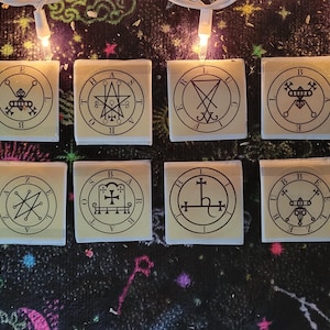 May include: Twelve square, cream-colored candles with black sigils and symbols are arranged on a dark, patterned surface. The candles are lit by a string of small, warm-toned lights, creating a mystical and esoteric aesthetic.