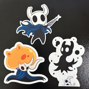 May include: Three die-cut stickers featuring characters from a video game. The top sticker shows a white character with a blue cape and a sword. The bottom left sticker has an orange and blue character. The bottom right sticker is a black and white character.